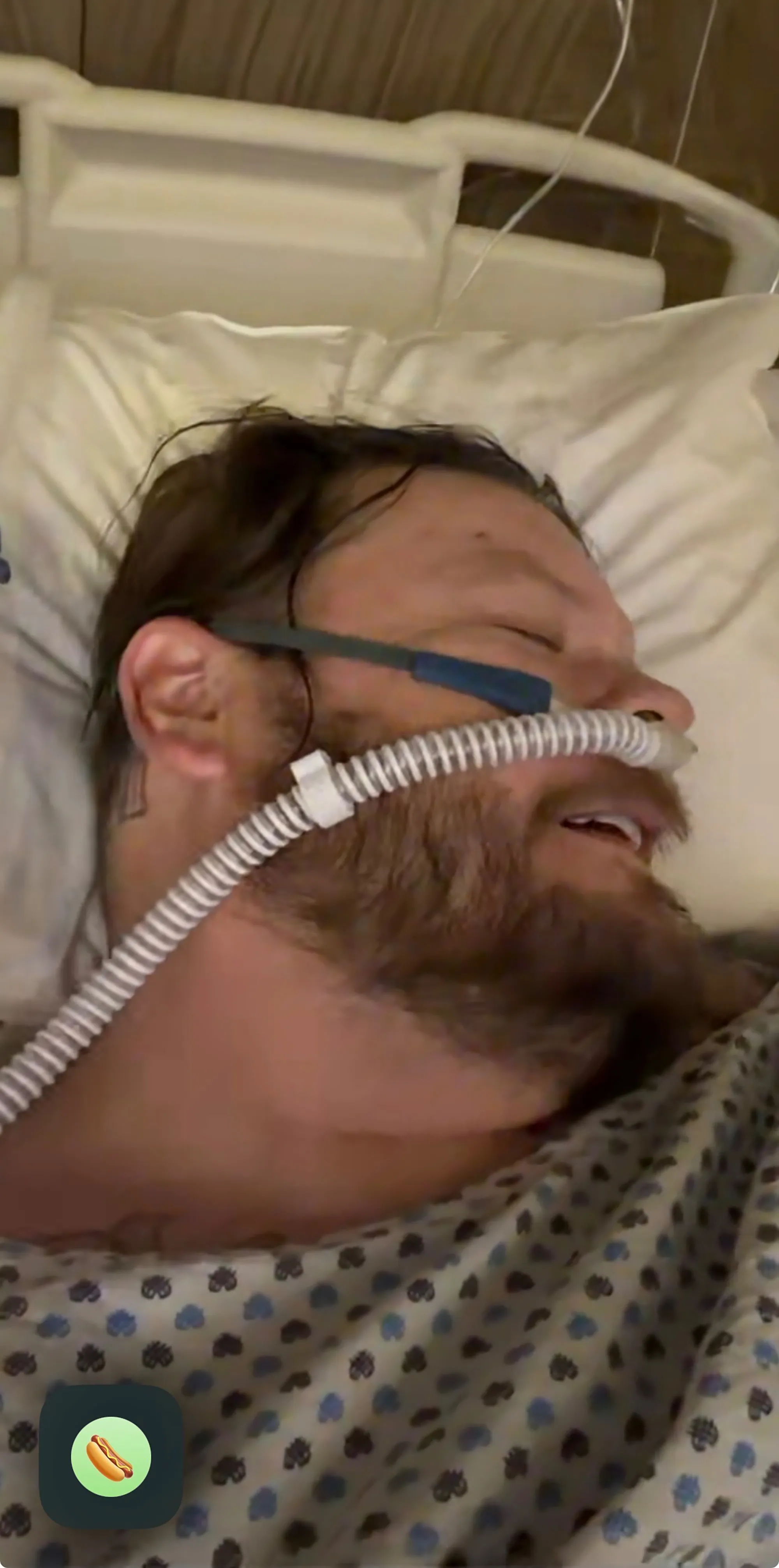 Corey Harrison in a hospital bed with a breathing tube, wearing a hospital gown.