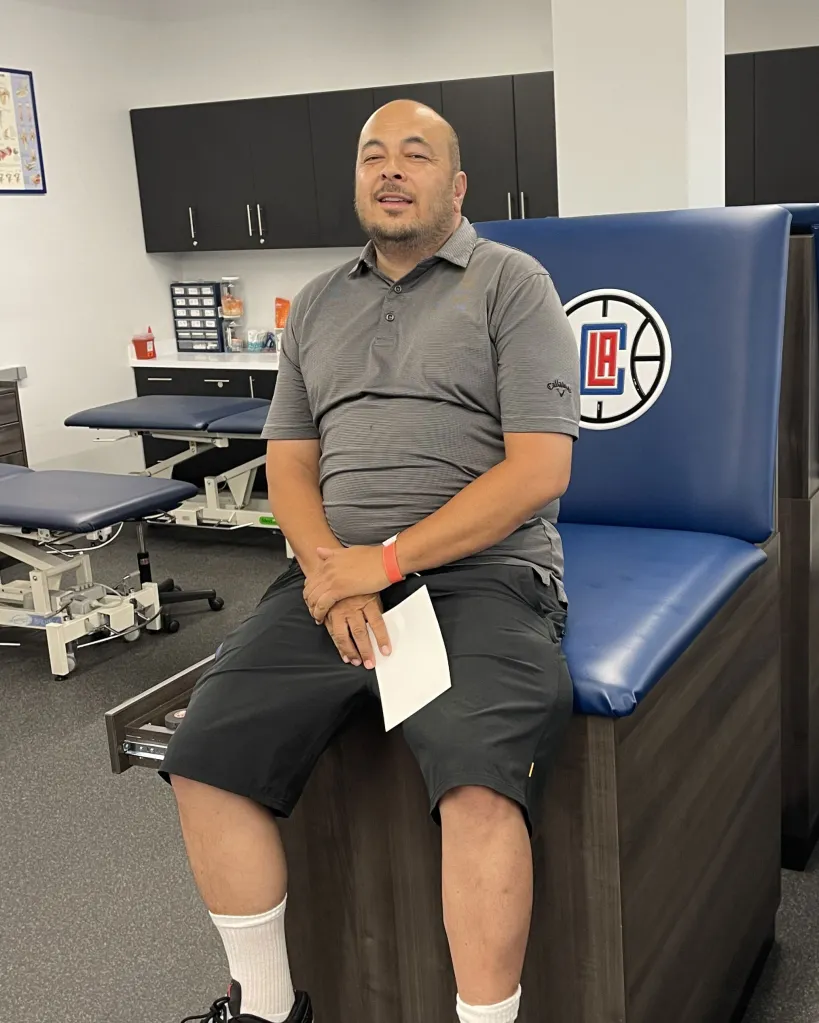 Rhett Pascual, a bald man with a beard, sitting on an examination table with his hands clasped and a paper resting on his lap.
