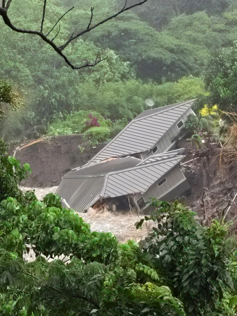 A house destroyed and partially submerged in floodwaters, with a large muddy embankment where the house once stood.