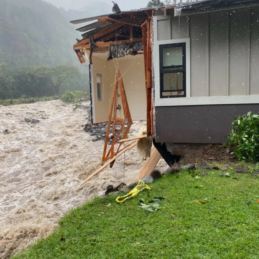 A house damaged by floodwaters with a raging brown river beside it.