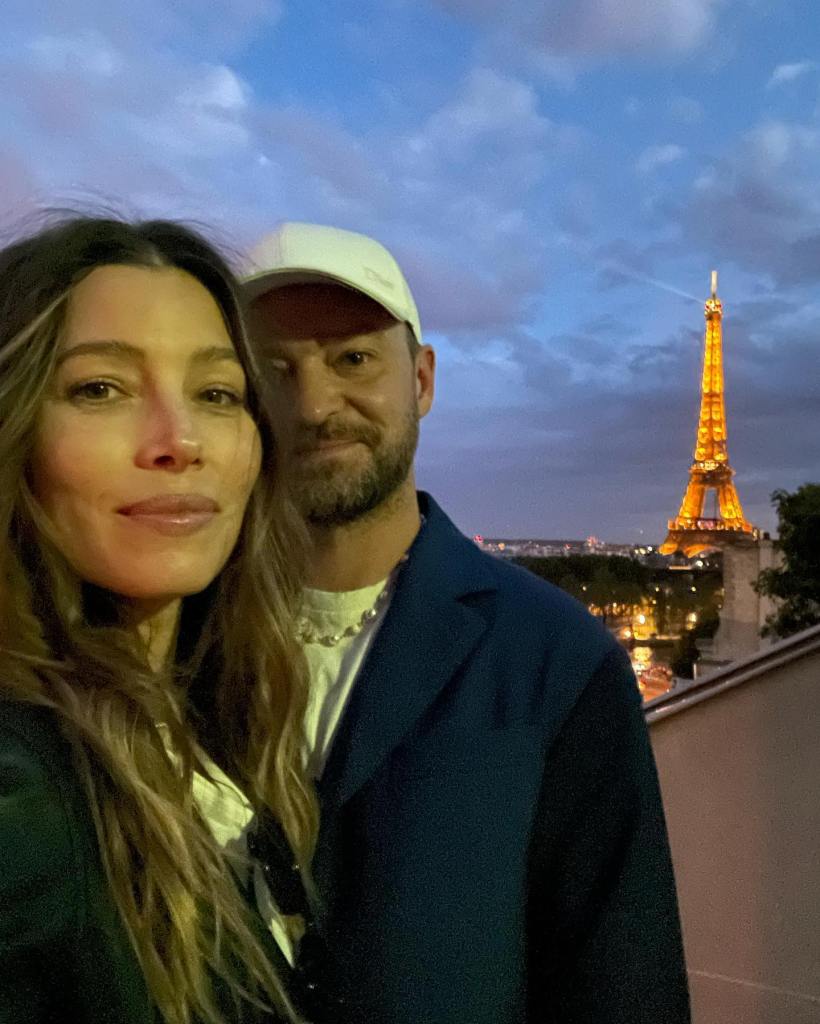 Jessica Biel and Justin Timberlake posing for a selfie in front of the illuminated Eiffel Tower at dusk.