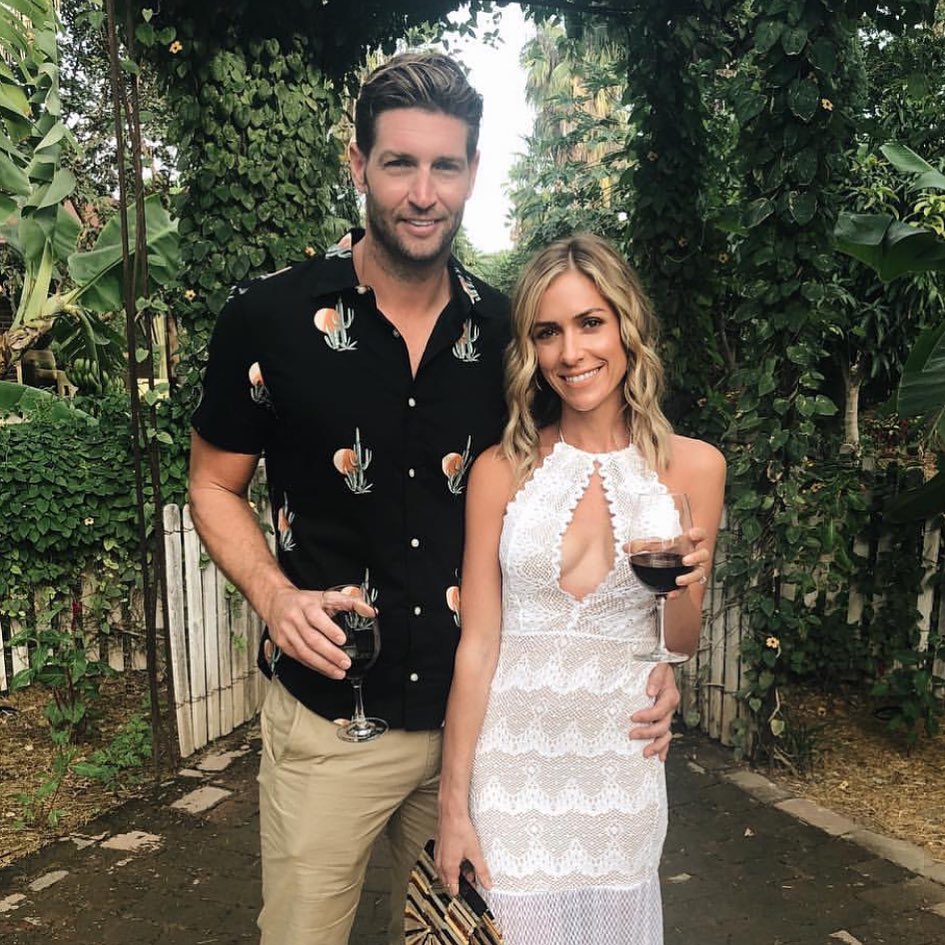 Kristin Cavallari and Jay Cutler pose with wine glasses under a leafy archway.