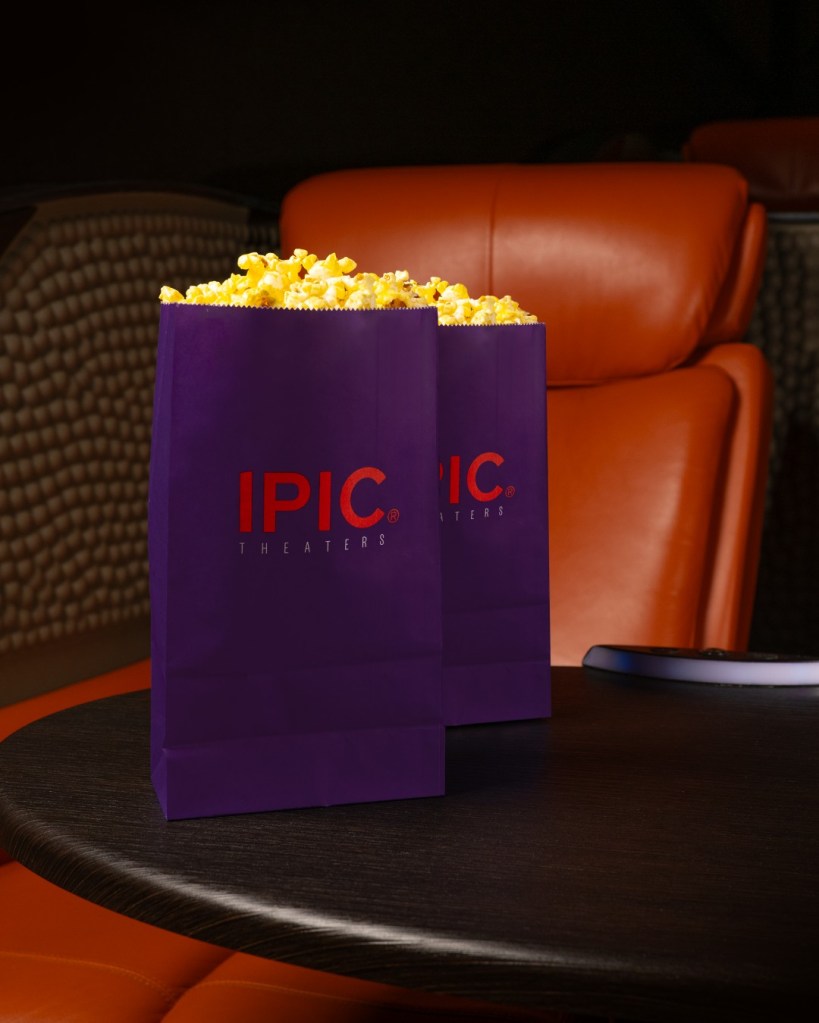 Two purple bags of IPIC Theaters popcorn sit on a dark wooden table in front of orange movie seats.