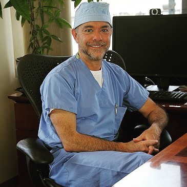 A smiling male doctor in blue scrubs and a surgical cap sits at a desk.