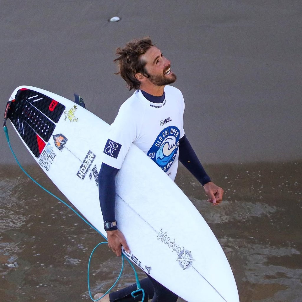 Lucas Filardi-Taub in a SLO CAL OPEN shirt carrying a surfboard on the beach.