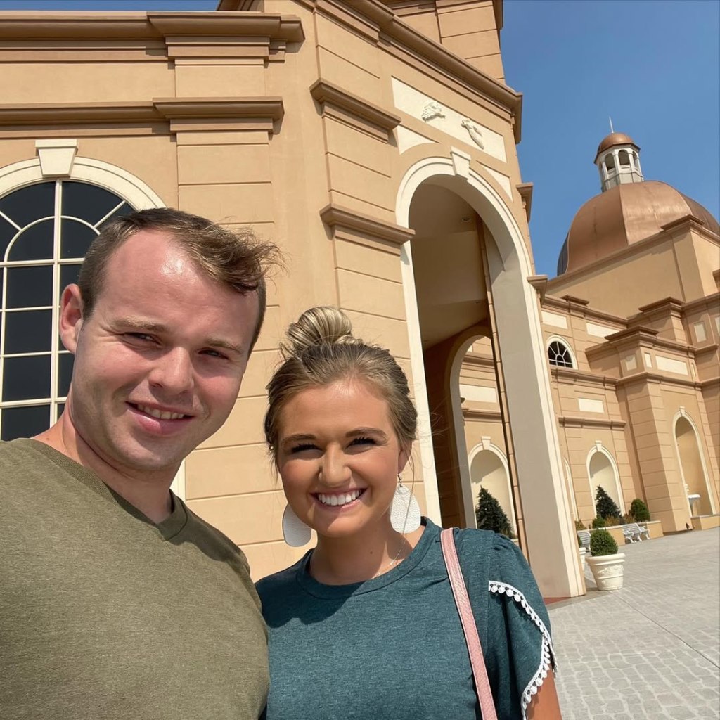 Joseph Garrett Duggar and his wife Kendra Caldwell Duggar smiling for a selfie in front of a large, tan building with a domed roof.