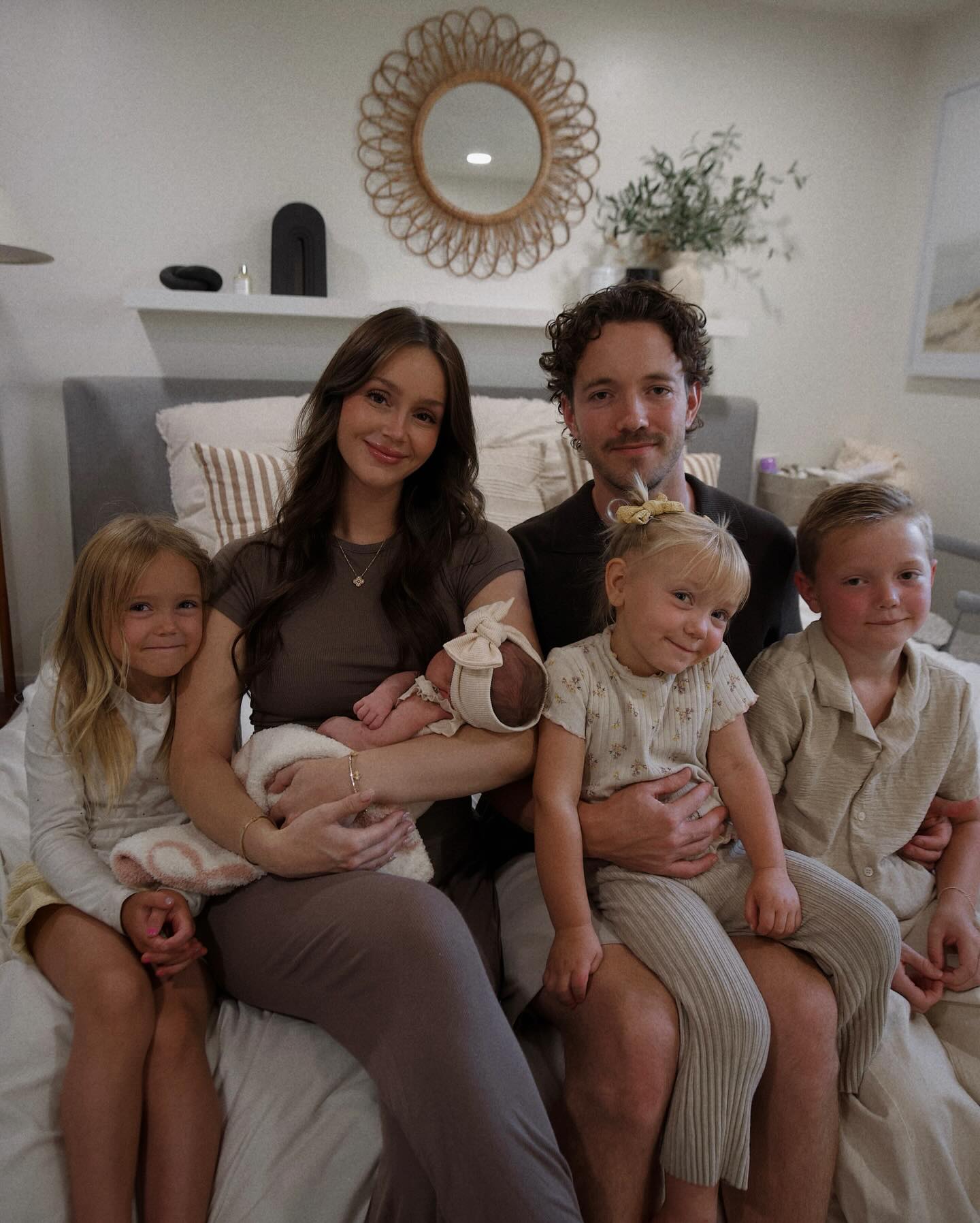 A family of six with four children, including a newborn, sitting on a bed.