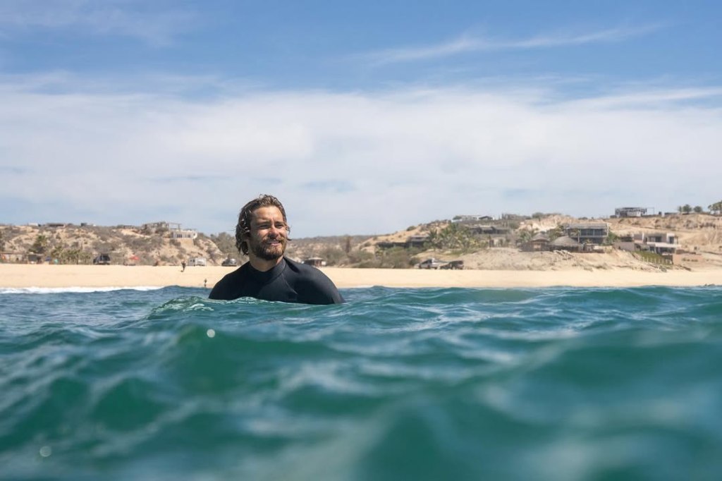 Lucas Filardi-Taub in the ocean, wearing a wetsuit with a sandy beach and hillside with buildings in the background.