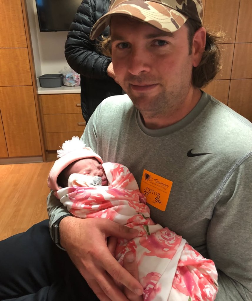 Jamie Watson camouflage hat holds a swaddled newborn in a hospital room.
