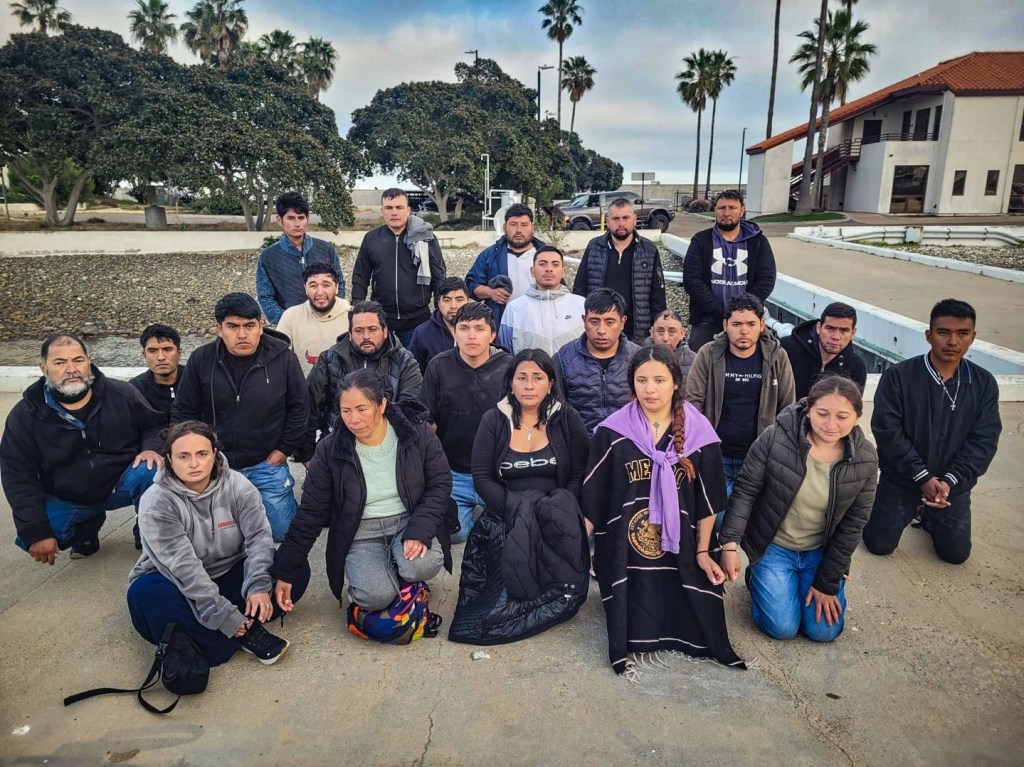 Twenty-three immigrants who were arrested by border authorities are pictured outside, some kneeling, with palm trees and a building in the background.