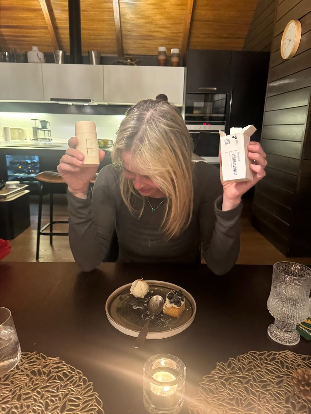 Chelsea Handler looking at two small boxes while seated at a table with dessert.