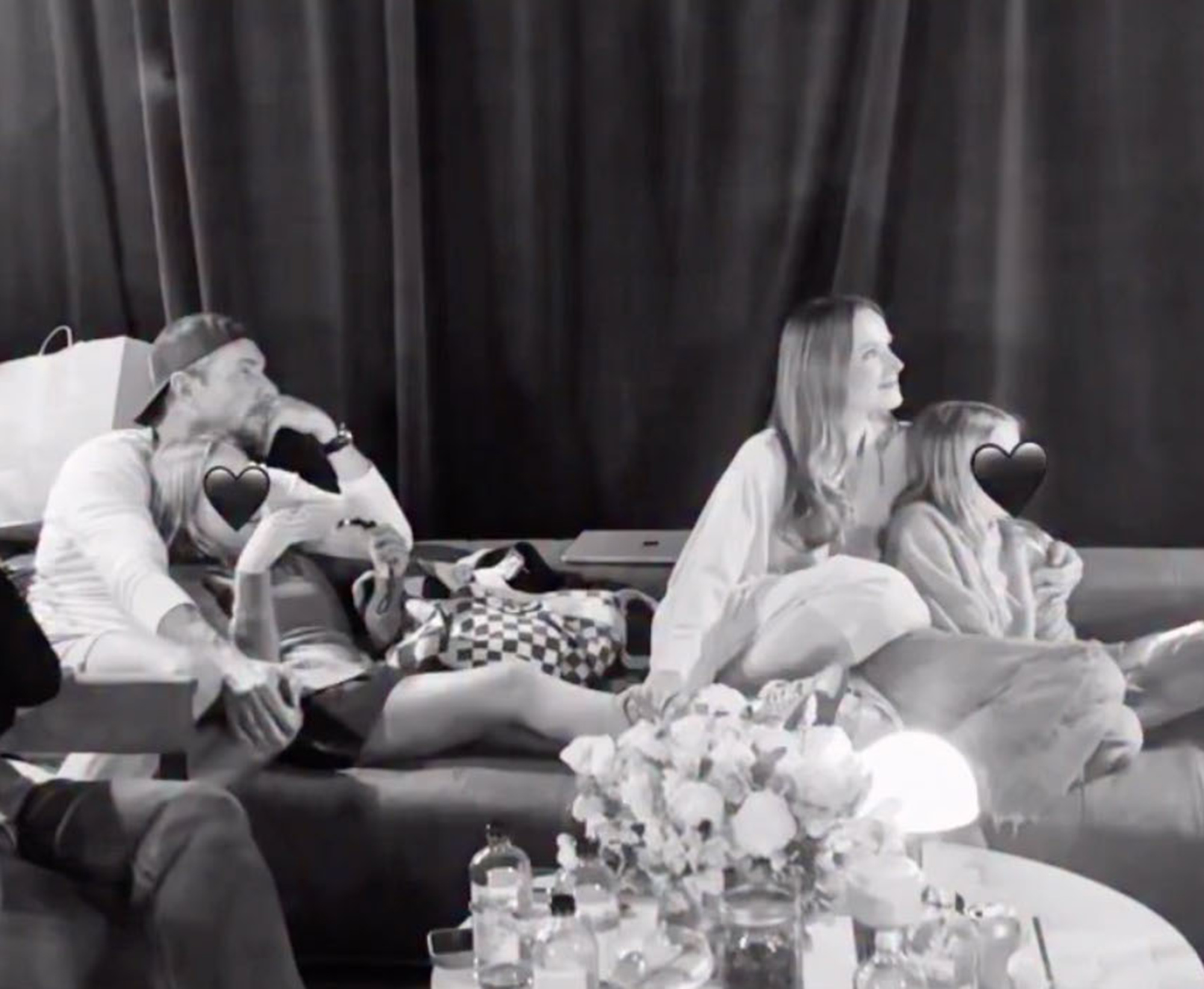 Black and white photo of Dax Shepard, Kristen Bell, and their two children relaxing on a couch, with their faces partially obscured by black heart emojis.