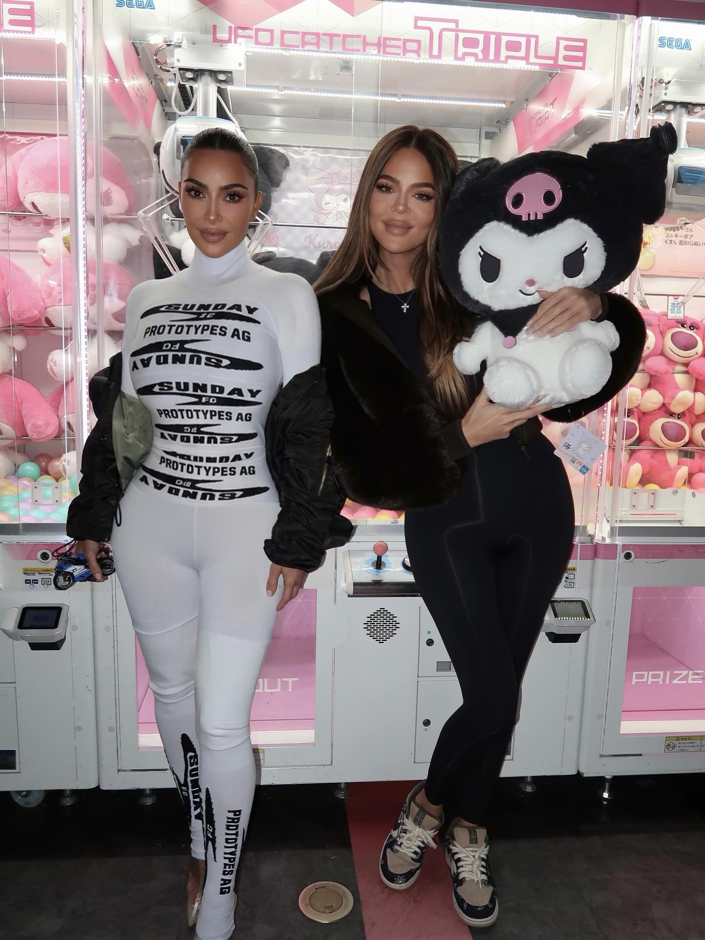 Kim Kardashian and Khloé Kardashian pose in front of claw machines.