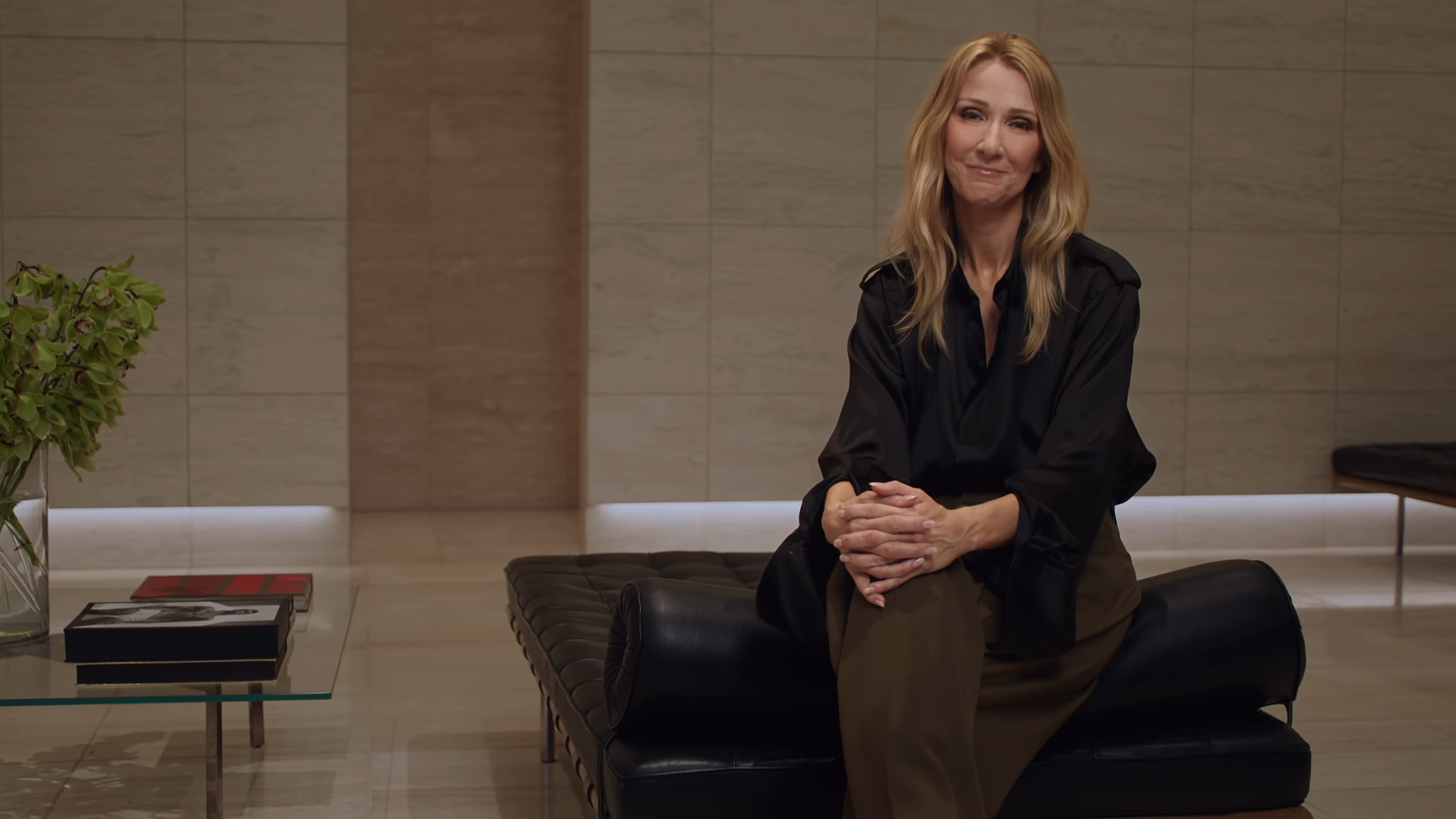 Celine Dion sits on a black leather bench with her hands clasped, smiling at the camera.