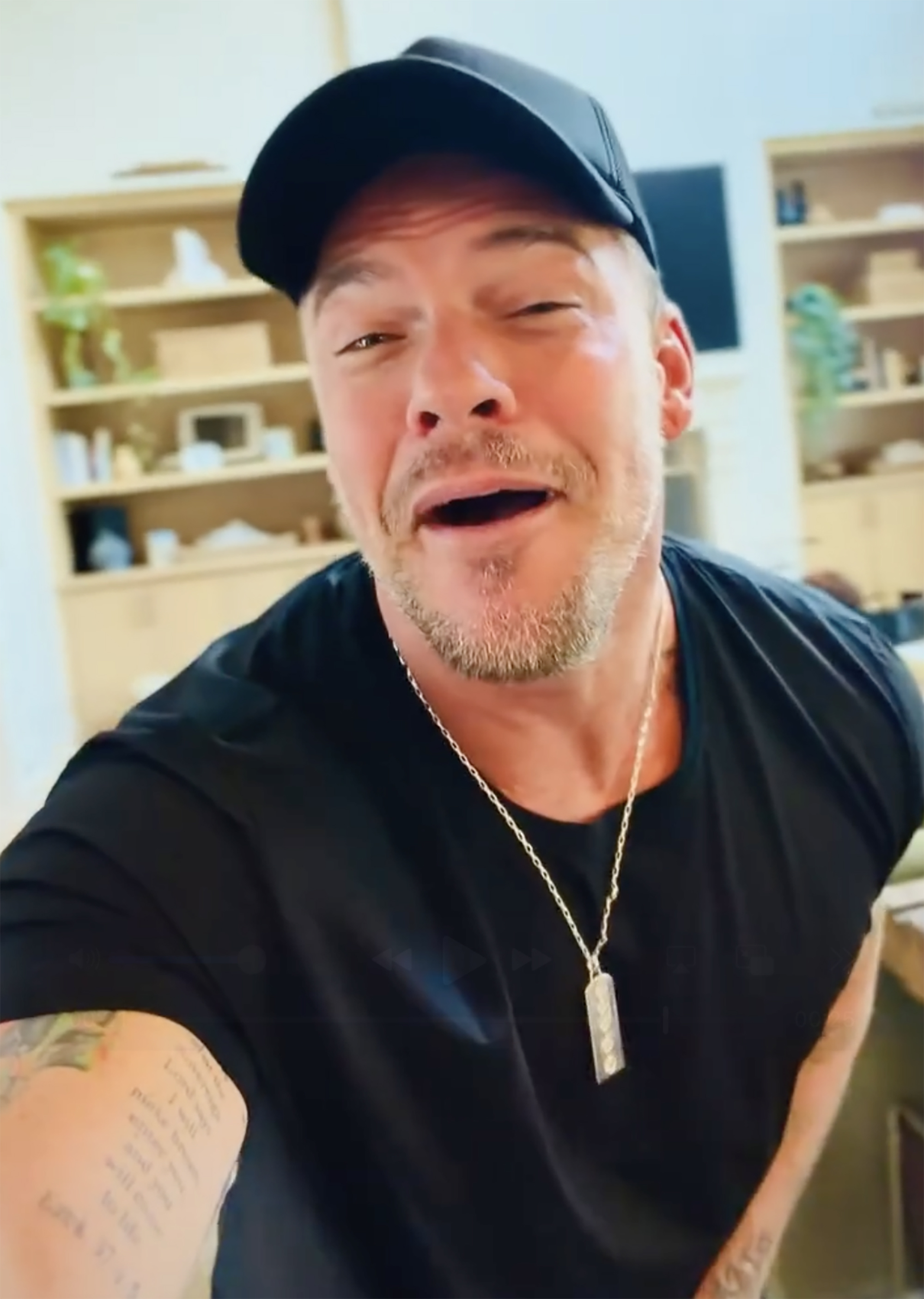 Alan Ritchson in a black cap and shirt, winking and smiling.