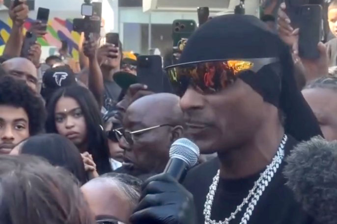 Snoop Dogg wearing a black du-rag and sunglasses, speaking into a microphone.