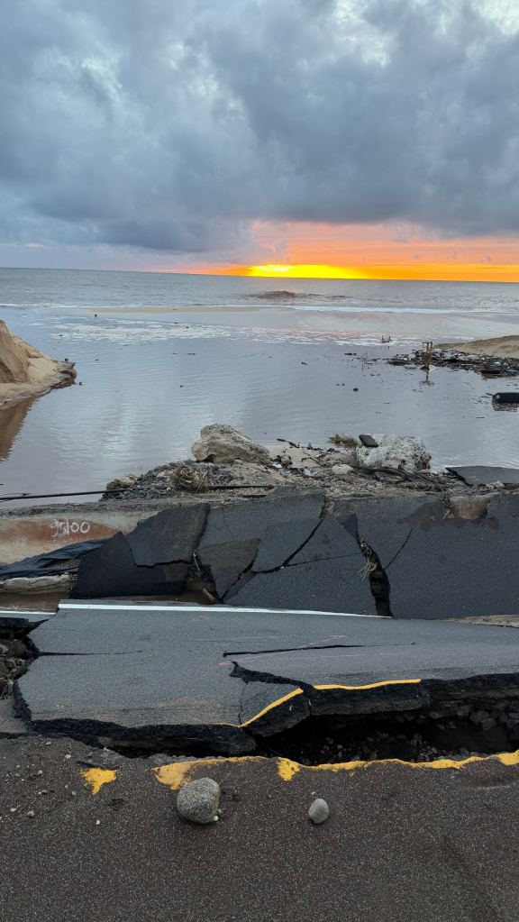 A damaged road with cracked asphalt, facing a beach and ocean at sunset, with debris scattered around.