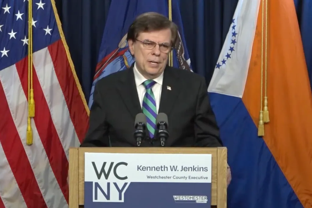 Councilman Francis Sheehan speaking at a podium with American and New York flags in the background.