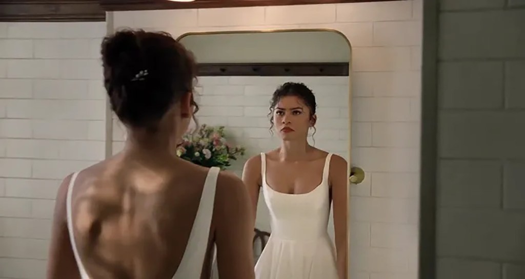 Zendaya looking in a mirror with a serious expression, wearing a white dress.