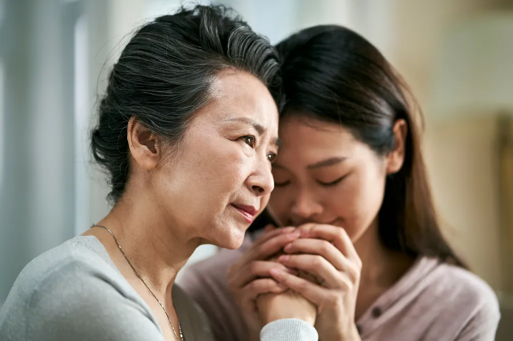 An adult daughter holding her mother's hands and resting her forehead on her mother's head, comforting her mother who is suffering from mental illness.