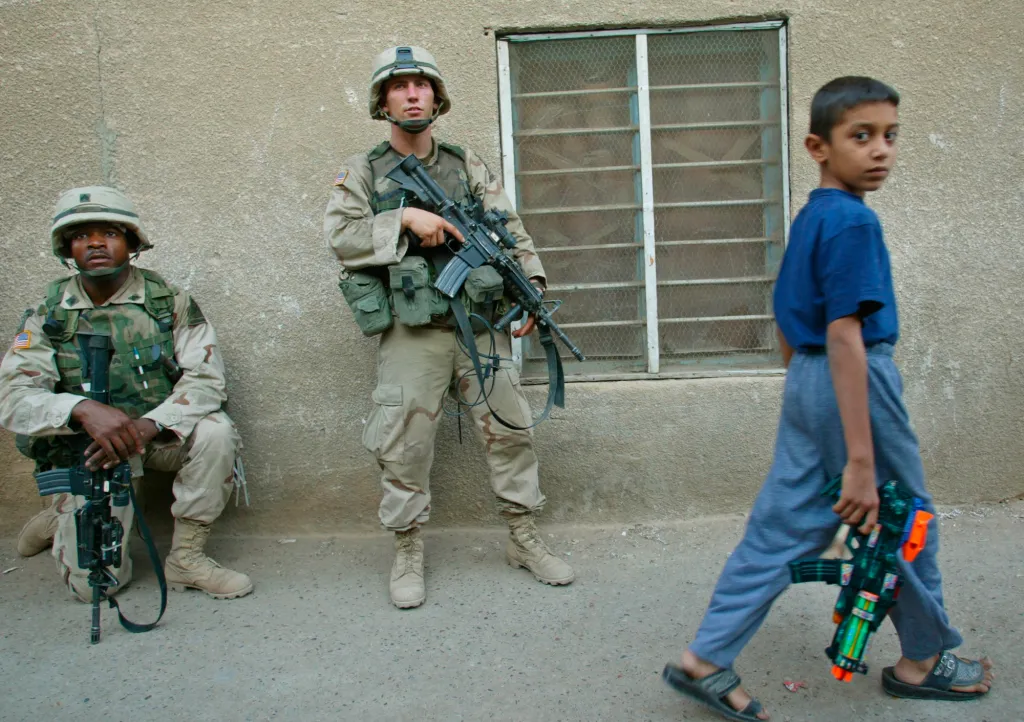 A boy with a toy gun walks past two armed US soldiers.