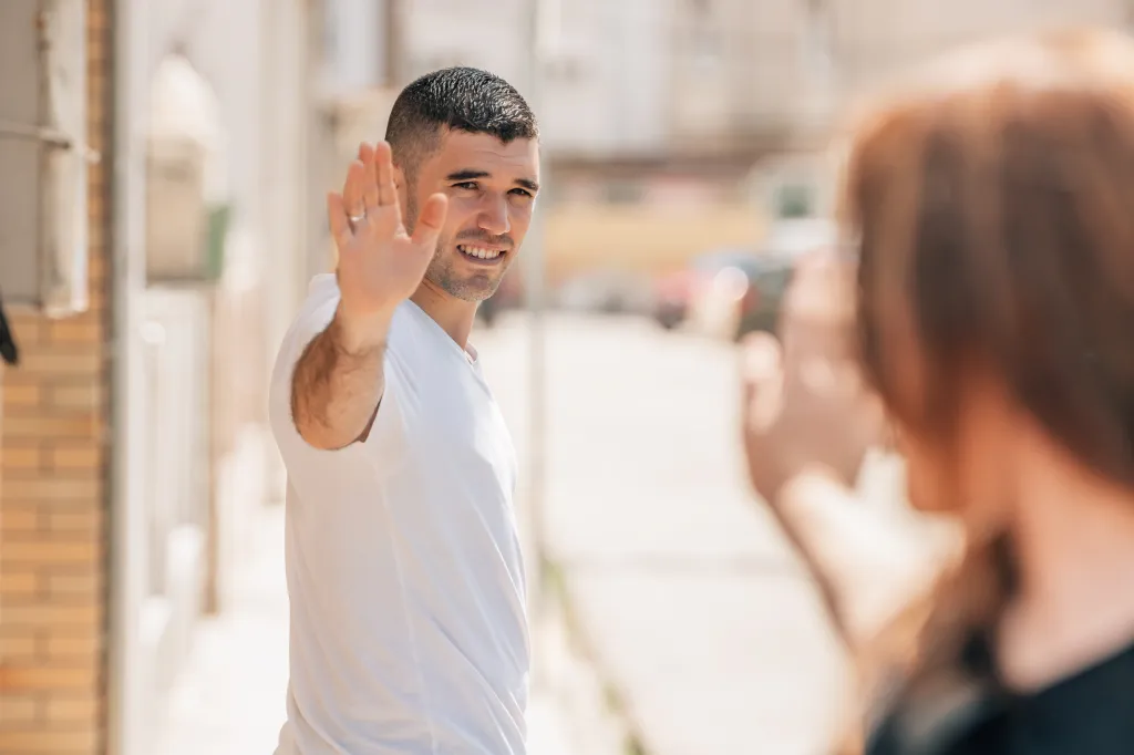 A man smiling and waving to a woman on a city street.