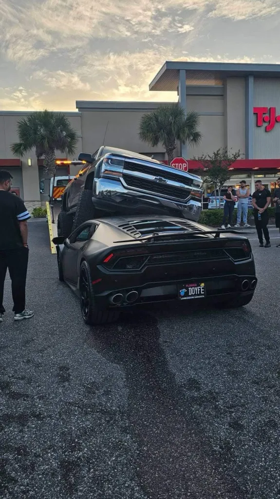 A black pickup truck sits on top of a black Lamborghini Huracan in a parking lot.
