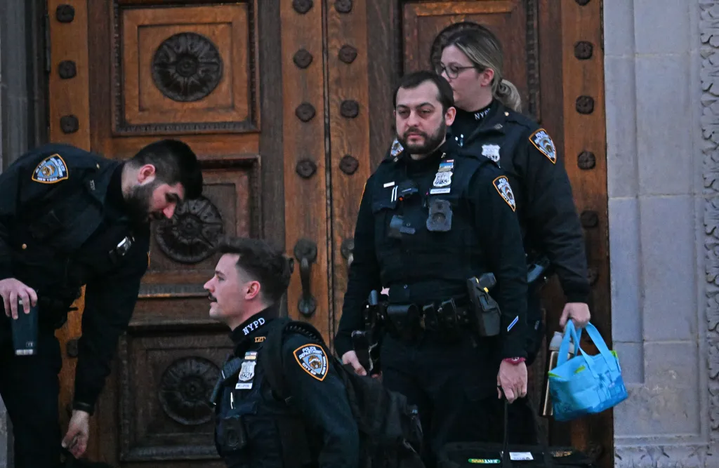 Four uniformed police officers stand in front of large wooden doors.