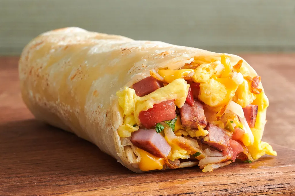 A breakfast burrito with eggs, ham, and cheese, visible from one open end, sits on a wooden surface.
