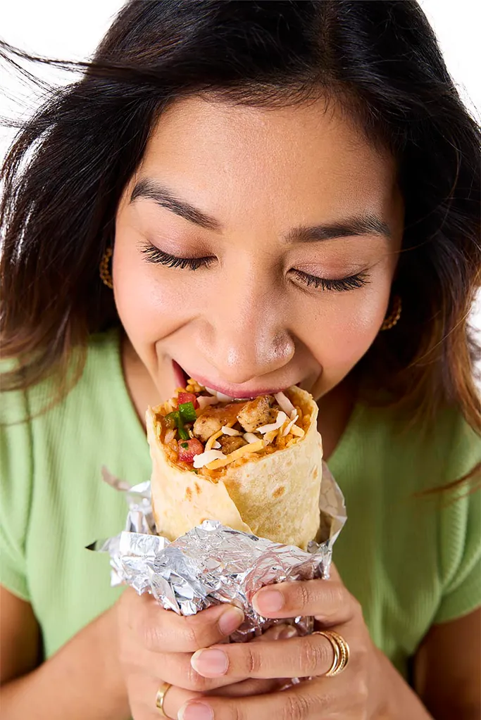 A woman with closed eyes taking a bite of a burrito wrapped in foil.