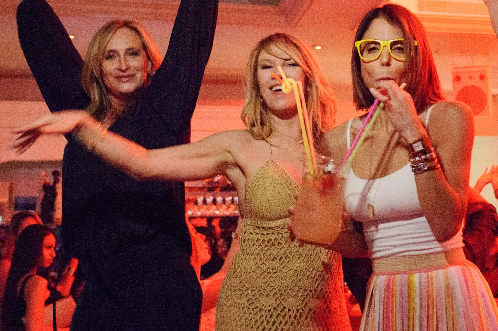 Three women dancing, two holding colorful drinks with multiple straws, one wearing yellow glasses.