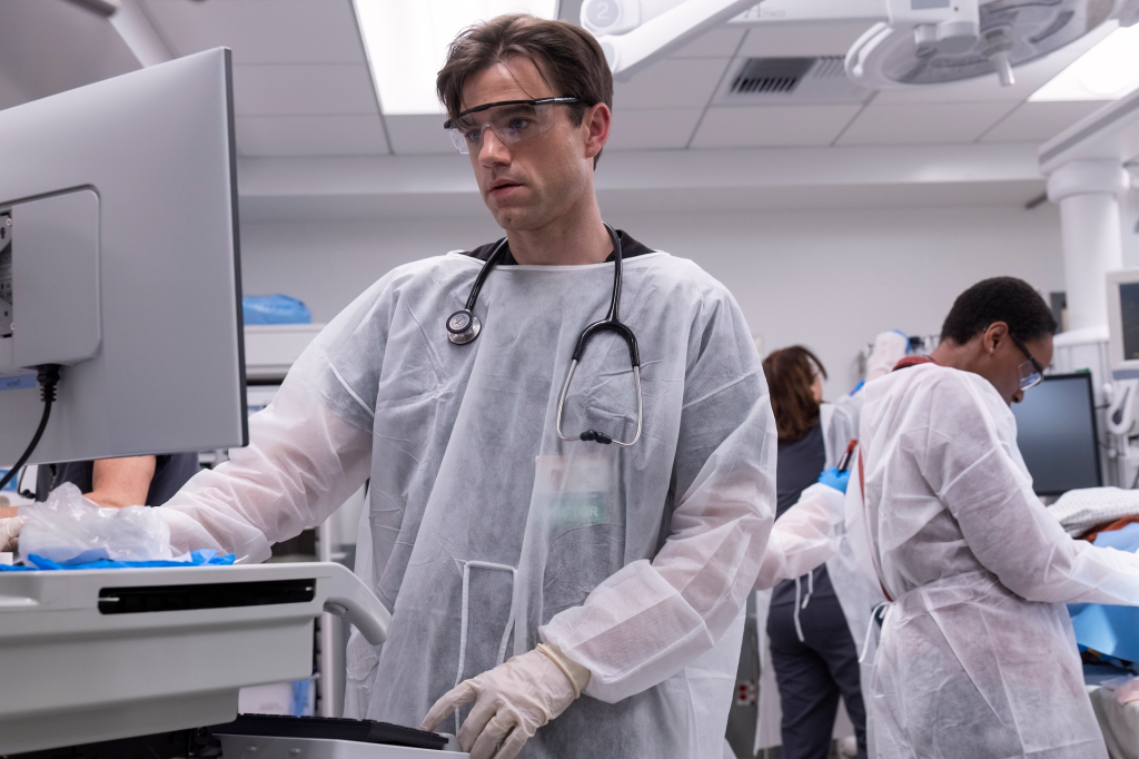 A doctor wearing protective gear and safety glasses reviews information on a computer monitor in a medical setting.
