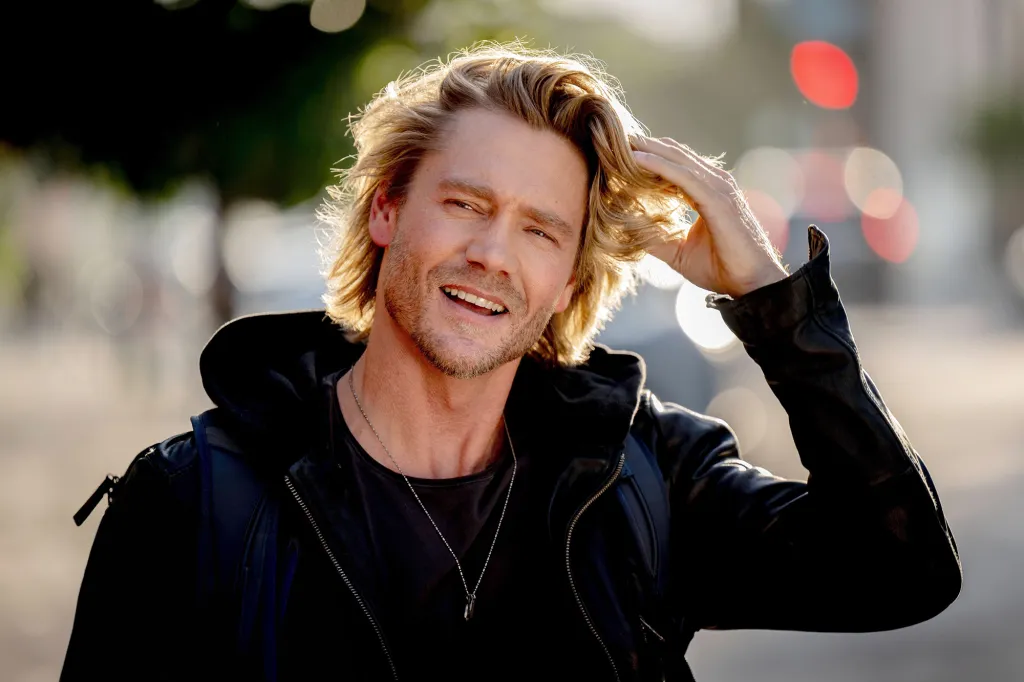 Man with blonde hair smiling, touching his hair with one hand.