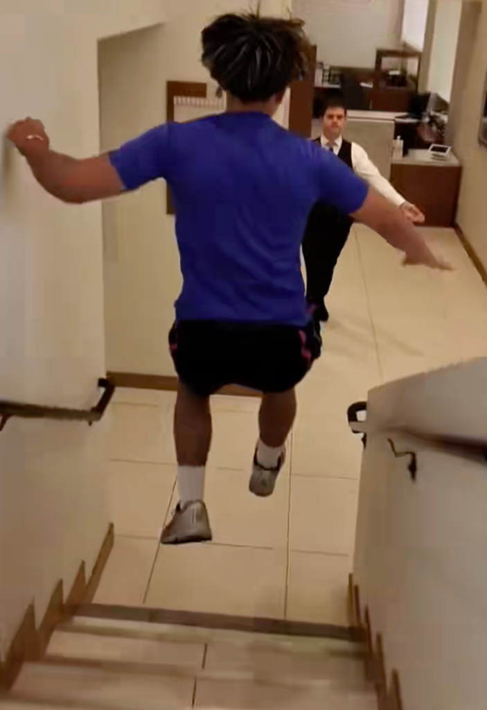 A man in a blue shirt leaps up stairs as a man in a black vest watches.