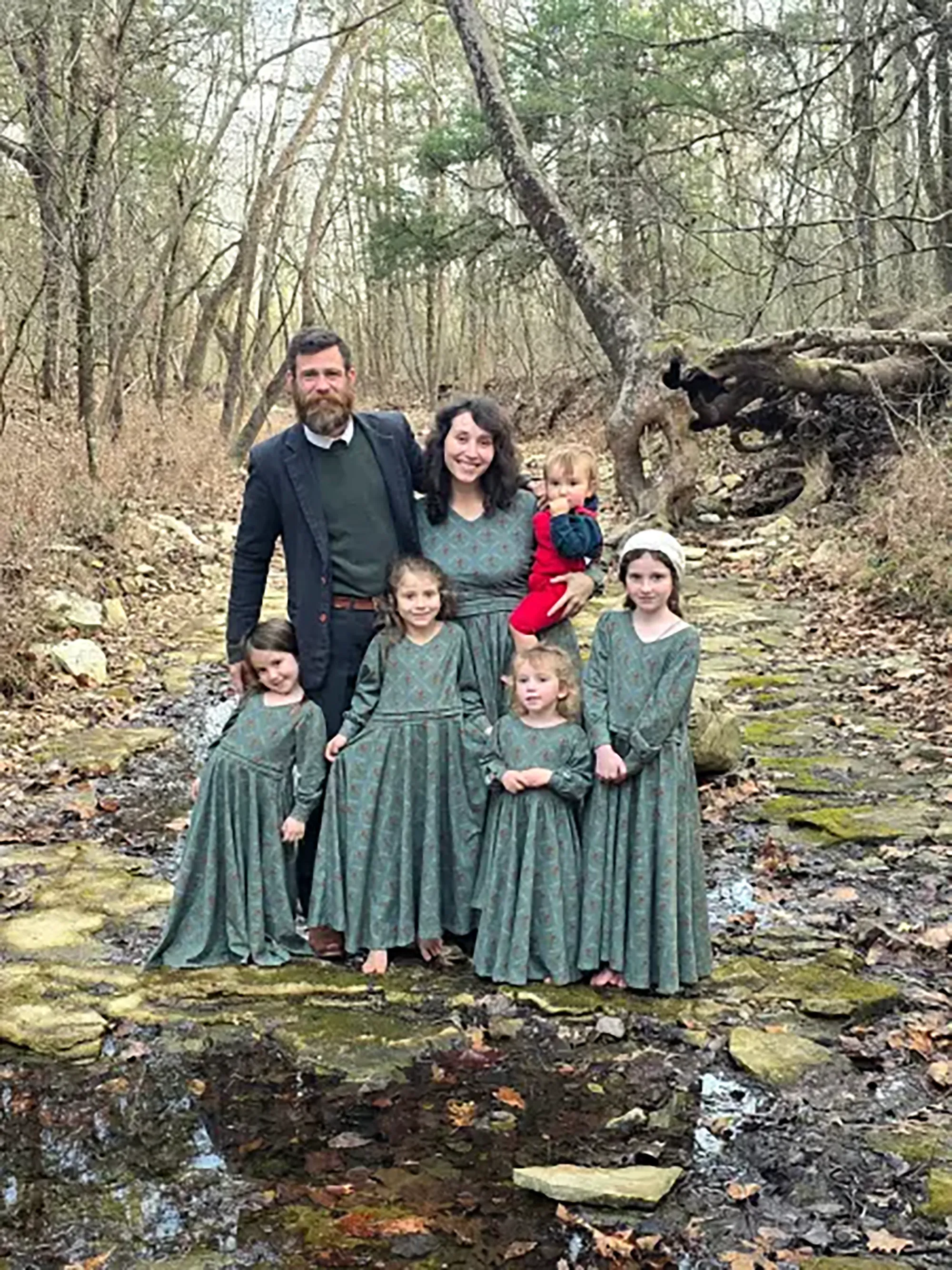 Husband, wife, and their five children stand in a wooded area with bare trees and a shallow stream.