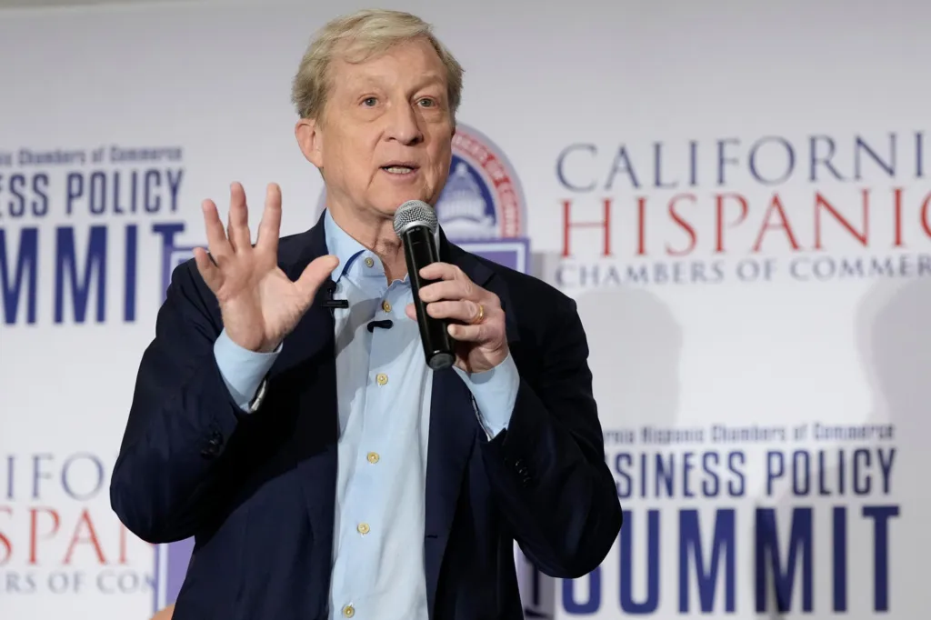 Tom Steyer speaking into a microphone with his hand raised at the California Hispanic Chambers of Commerce Business Policy Summit.