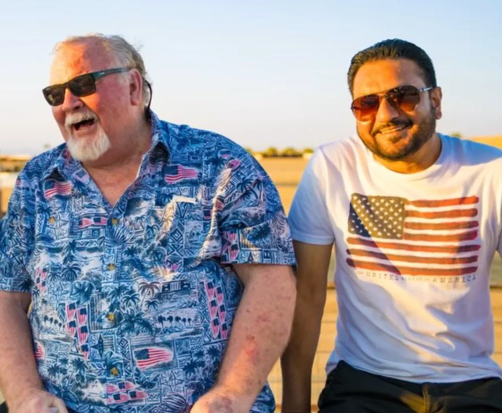 Two men smiling, one in a blue Hawaiian shirt with American flags and the other in a white t-shirt with an American flag.