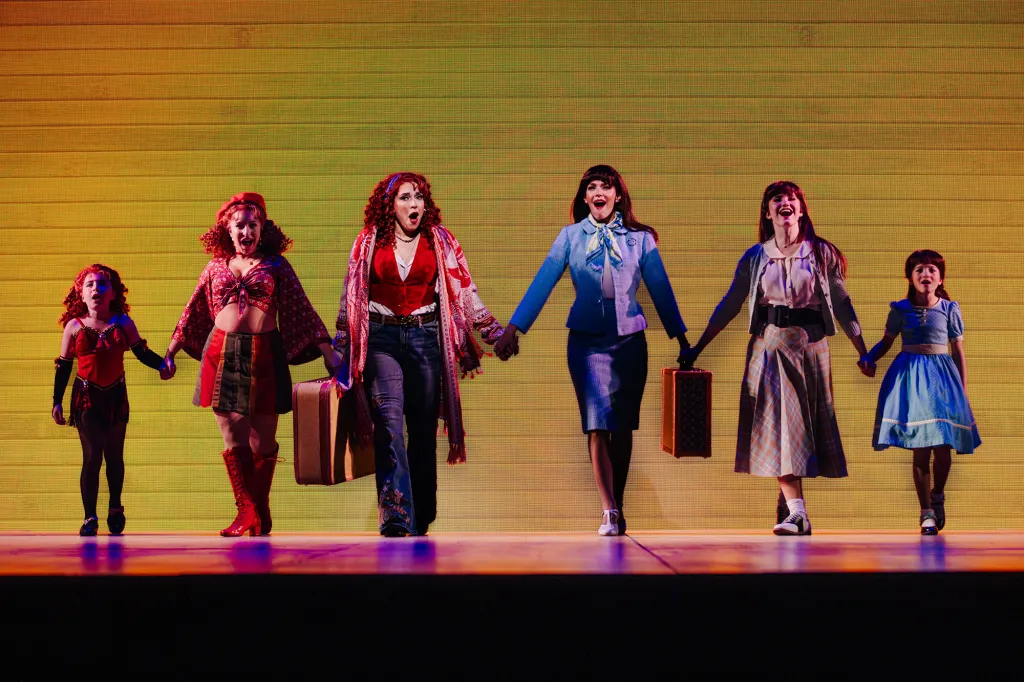 Six women and girls in varying costumes and carrying suitcases hold hands and sing on stage.