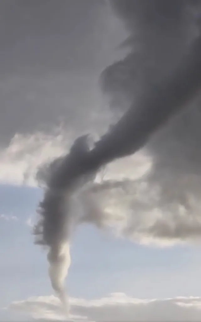 A dark grey tornado extending from thick clouds toward the bottom of the frame against a lighter sky.