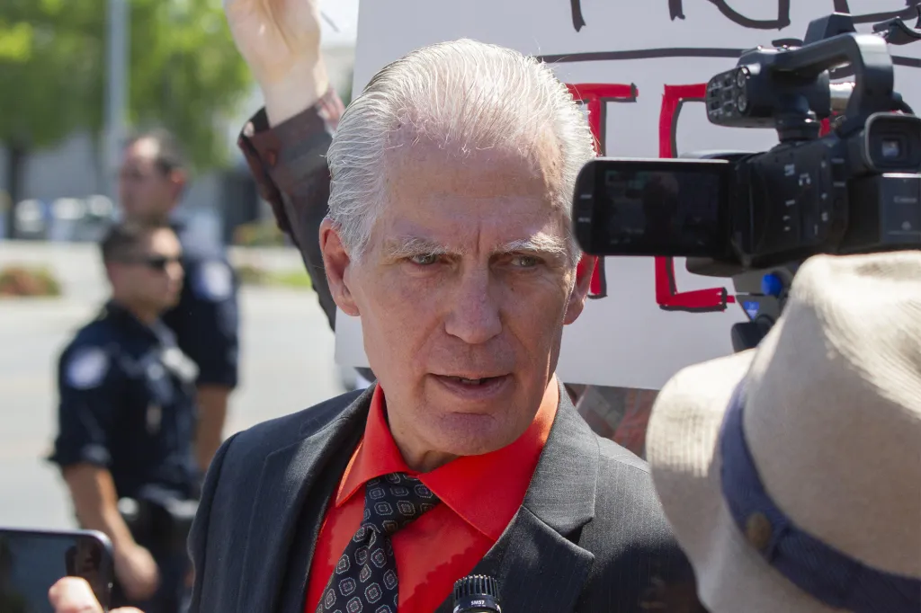 A man in a red shirt and black suit jacket with gray hair and green eyes speaks into a microphone, with a camera and person in a hat to his right, and two police officers in the background.
