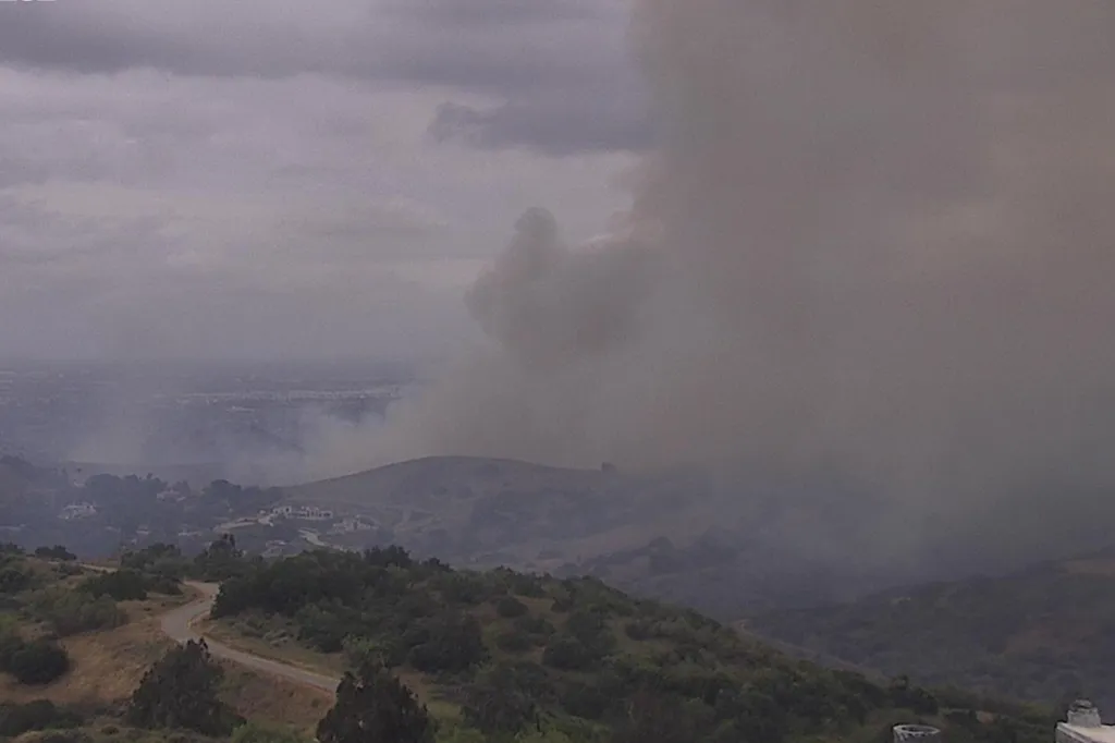 Large plume of smoke rising from a hillside fire, obscuring the sky and distant buildings.