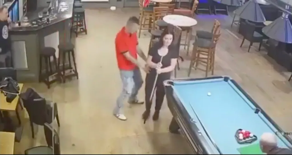 A man in a red shirt grabbing a woman holding a pool cue in a bar.