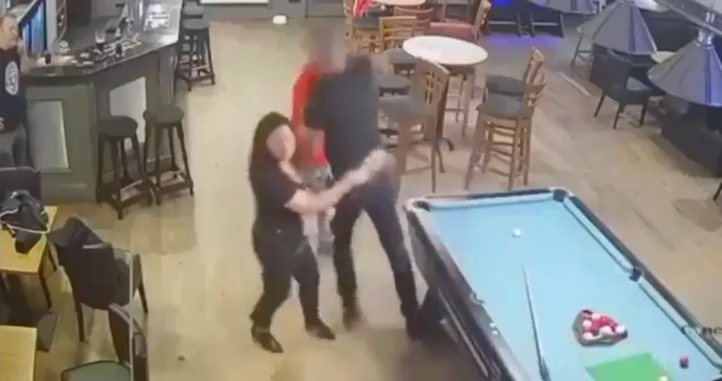 A woman attempts to break up a fight between two men in a bar, near a pool table.