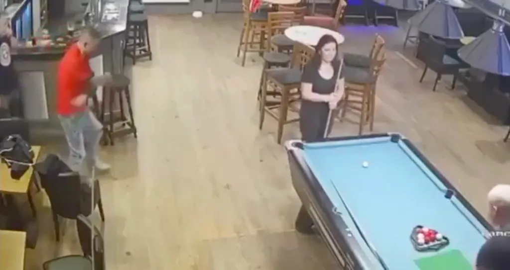 A man in a red shirt and a woman in a black shirt playing pool in a bar.
