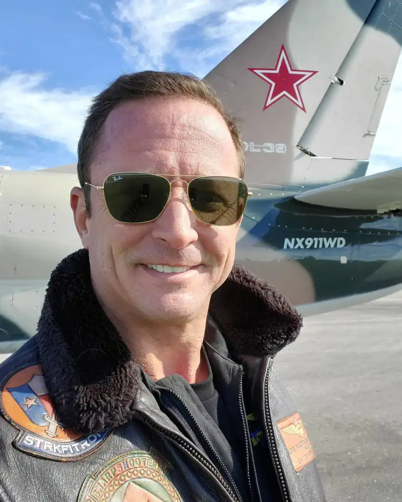 A man in sunglasses and a bomber jacket smiling in front of an L-39 jet with a red star on the tail.