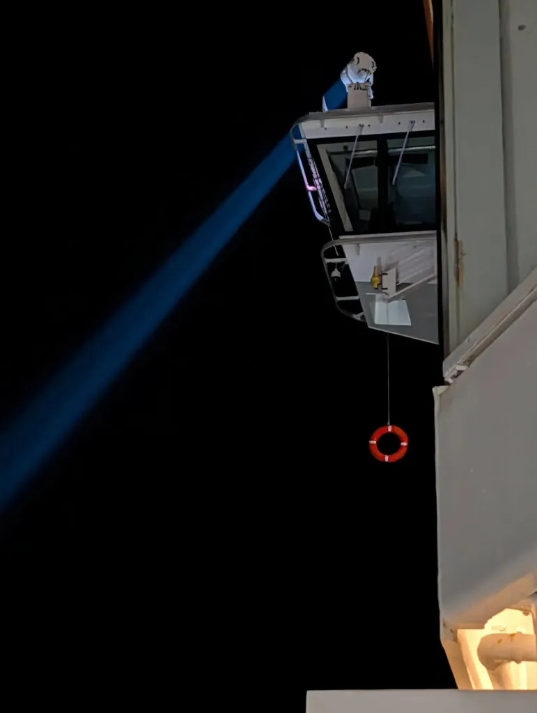 A cruise ship at night with a searchlight beam shining into the dark water, and a life buoy hanging from the side of the ship.