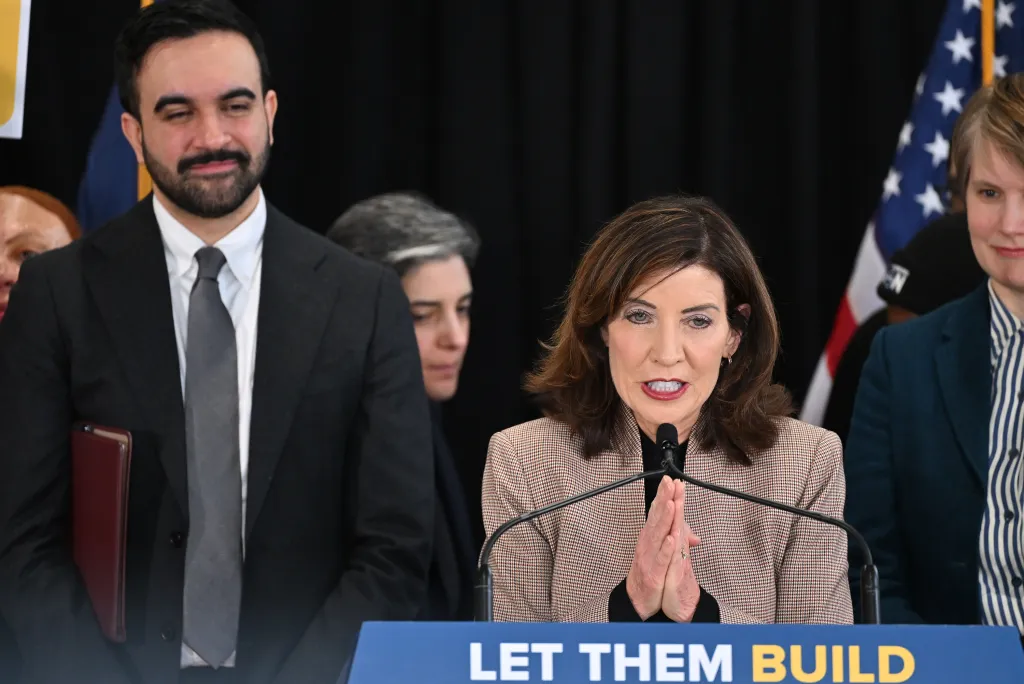 NYS Governor Kathy Hochul speaks at a podium with NYC Mayor Zorhan Mamdani standing nearby in Brooklyn.