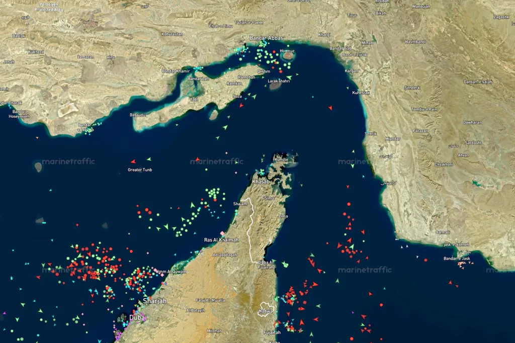 Satellite view of marine traffic in the Strait of Hormuz showing clusters of green, red, and light blue vessels.