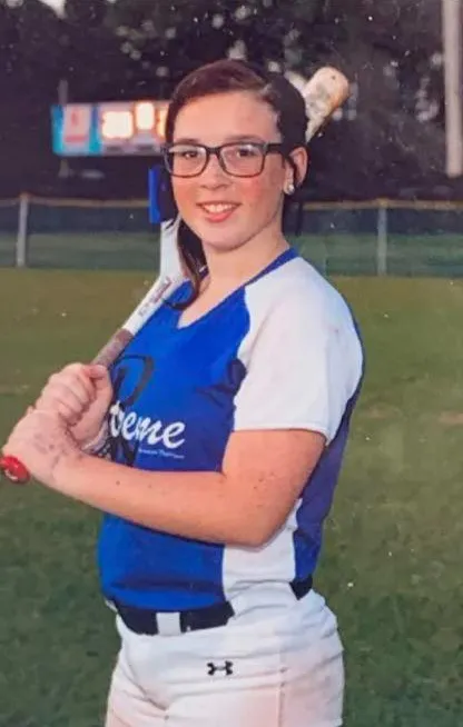 Keziah Arionna Luker, a young woman wearing glasses and a baseball uniform, holding a bat and smiling.
