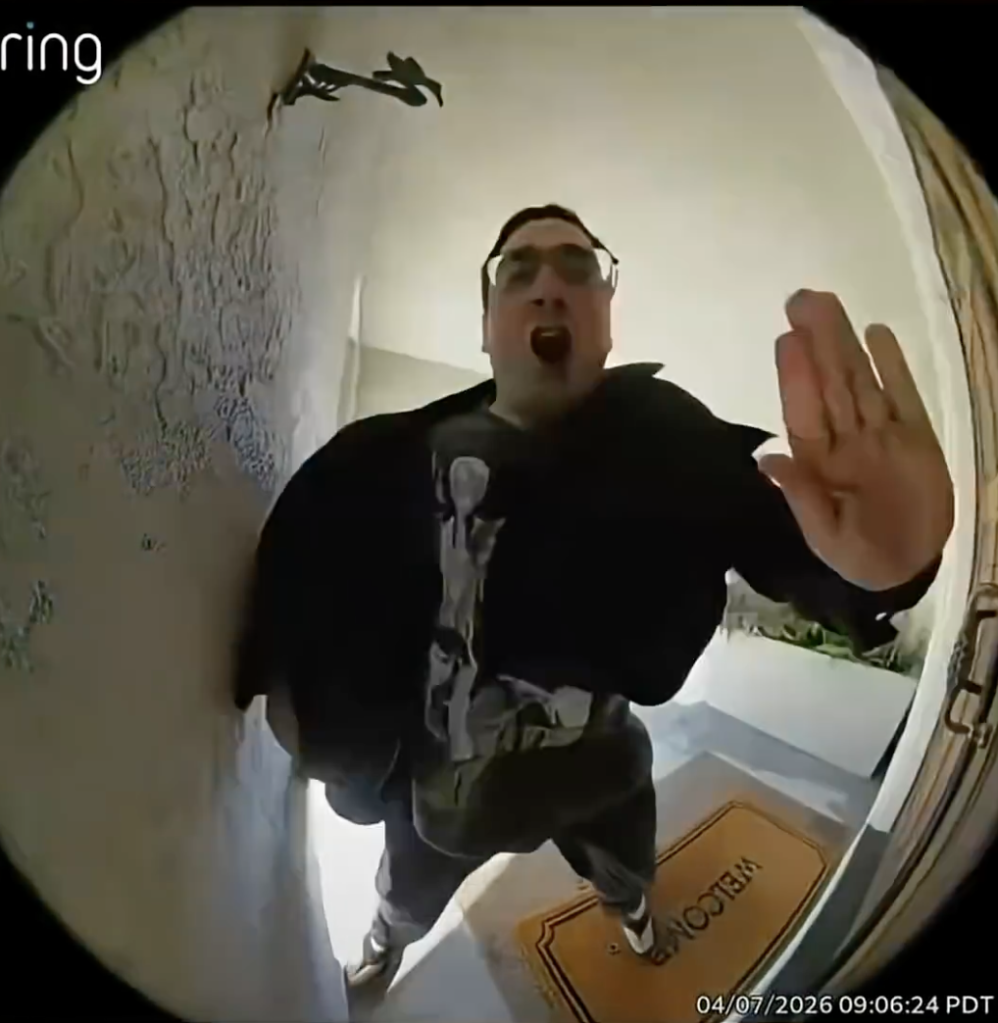A man with glasses and a black jacket, captured by a Ring doorbell camera, standing on a